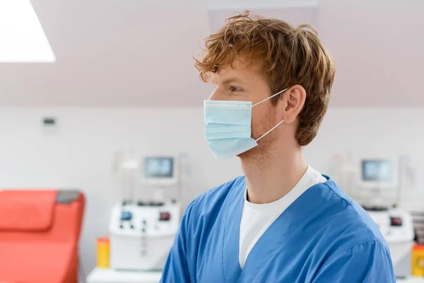 profile of redhead, young and positive doctor in medical mask and blue uniform looking away in blood donation center near transfusion machines with monitors on blurred background