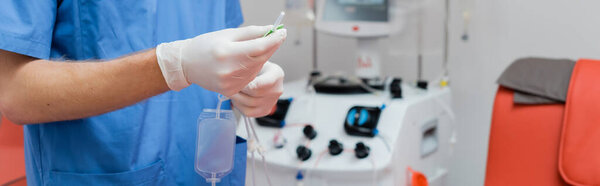 partial view of practitioner in blue uniform and latex gloves holding blood transfusion set near automated equipment and blurred near medical chair on blurred background in clinic, banner