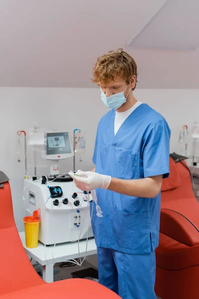 在献血中心的自动化设备和医疗椅子附近 戴着医疗面罩 乳胶手套和蓝色制服的红头发和年轻从业者手持输血器械 — 图库照片