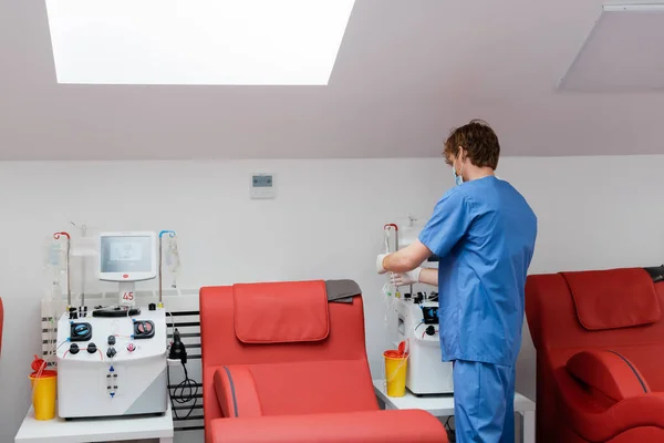 自動輸血機の近くの青の制服を着た若い赤毛の医者のバックビュー プラスチックカップ ドリップスタンド 献血センターの快適な医療椅子 — ストック写真