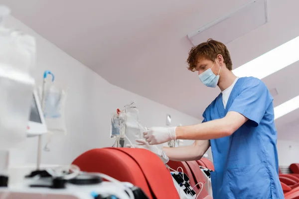 医療用マスクとラテックス手袋の赤毛の医療従事者は献血センターの自動化機器や医療椅子の近くに輸血セットを組み立てる — ストック写真