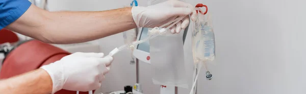 輸血セットと輸液袋を組み立てラテックス手袋の医師の部分的なビューは 献血センター バナーの滅菌環境で赤い医療椅子の近くの点滴スタンドに — ストック写真
