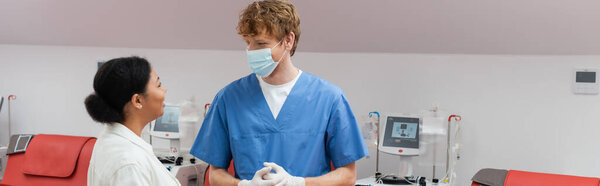 рыжеволосый врач в медицинской маске, латексные перчатки и синяя униформа беседуют с многорасовой женщиной возле автоматизированных аппаратов для переливания крови, капельниц и медицинских стульев в центре донорства крови, баннер
