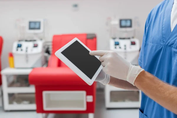 ラテックス手袋の医者の部分的なビューは 診療所でぼやけた背景にぼやけた医療椅子と輸血機の近くに空白の画面を持つデジタルタブレットを指しています — ストック写真