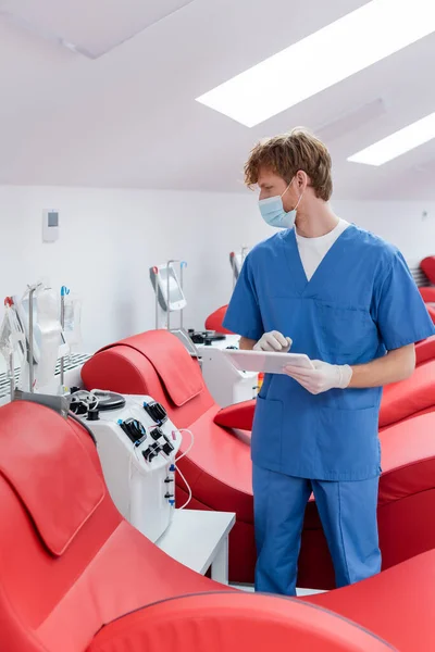 在献血中心 戴着医疗面罩的红头发医生拿着数码平板电脑 站在人体工学椅子和自动输血机旁边看着滴水 — 图库照片