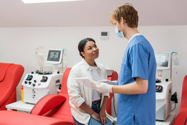 рыжеволосый врач в медицинской маске, синей униформе и латексных перчатках с цифровым планшетом и беседой с многорасовым донором, сидящим на эргономичном стуле возле аппаратов для переливания крови в клинике