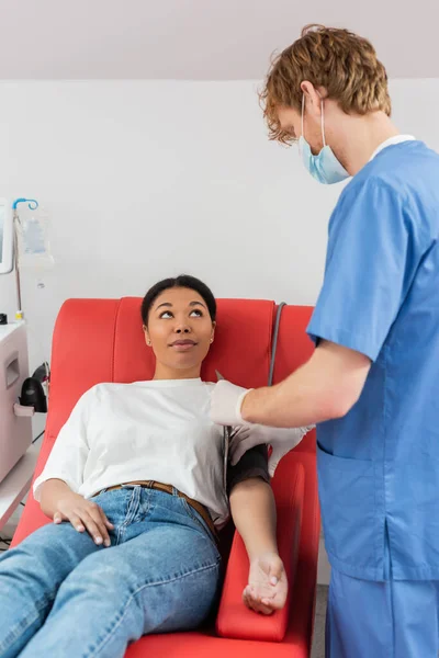 身穿医疗面罩 蓝色制服和乳胶手套的红头发医护人员将血压手铐戴在多种族女性的臂膀上 坐在实验室的医疗椅上 — 图库照片