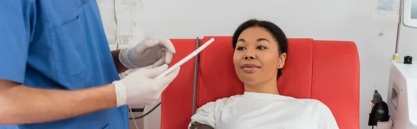 献血センター バナー 患者の医療椅子に座っている多人種の女性の近くのデジタルタブレットを使用して青い制服 医療用マスク ラテックス手袋の医療従事者 — ストック写真