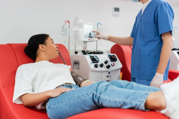 在医疗实验室 身穿蓝色制服和乳胶手套的医护人员在多种族妇女献血附近操作自动输血机 — 图库照片