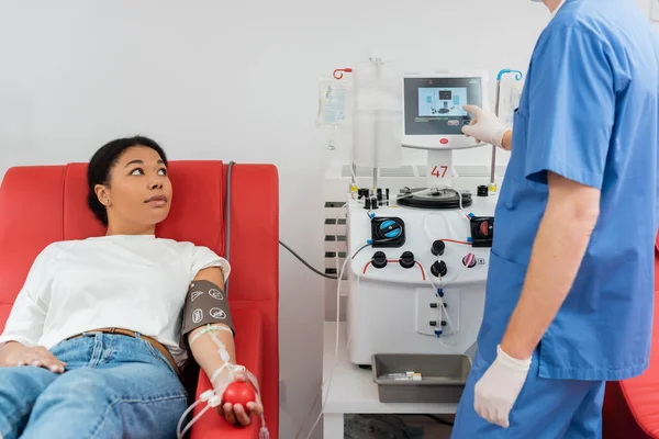 身穿蓝色制服和乳胶手套的医生在实验室献血时 接近多种族妇女坐在医用椅子上 手握橡皮球 操作输血机 — 图库照片