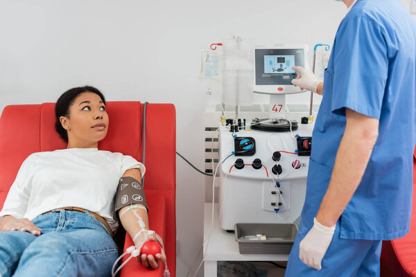 doctor in blue uniform and latex gloves operating transfusion machine near multiracial woman sitting on medical chair with rubber ball while donating blood in laboratory