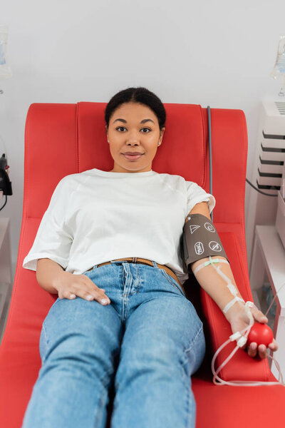 positive multiracial woman with pressure cuff, transfusion set and medical rubber ball sitting on medical chair and looking at camera during blood donation in clinic