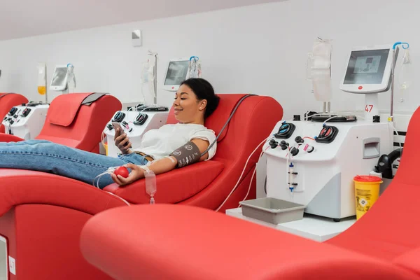 在诊所献血时 坐在自动设备旁边的医疗椅子上 一边笑一边拿着橡皮球和输血的多种族女人一边在智能手机上浏览互联网 — 图库照片