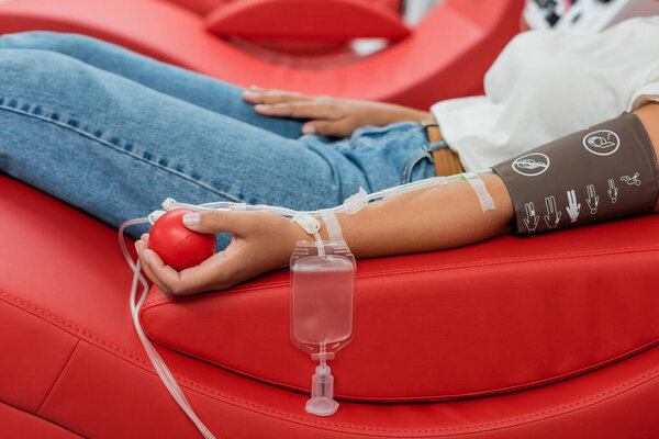 частичный взгляд на многорасовую женщину с набором для переливания крови и медицинским резиновым мячом, сидящим на эргономичном медицинском стуле во время сдачи крови в лаборатории