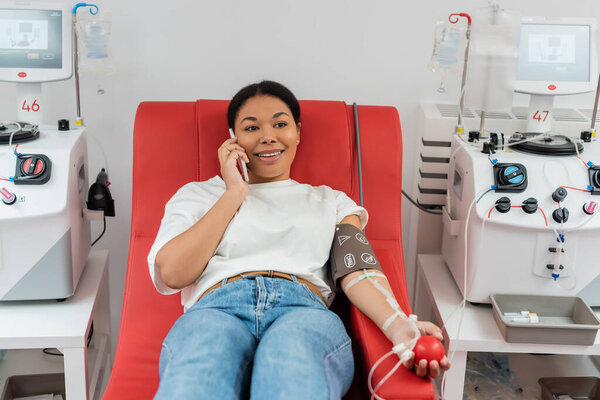 smiling multiracial woman with pressure cuff and rubber ball sitting on medical chair near transfusion machines and talking on mobile phone during blood donation in clinic