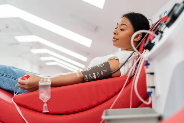 stock image side view of multiracial woman in pressure cuff and transfusion set holding rubber ball while sitting on comfortable medical chair near blurred transfusion machine in clinic