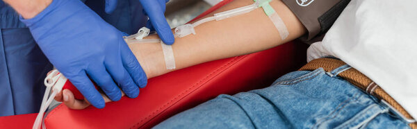 partial view of healthcare worker in latex gloves sticking band-aid on arm of multiracial woman sitting on medical chair with blood transfusion set, medical procedure, banner