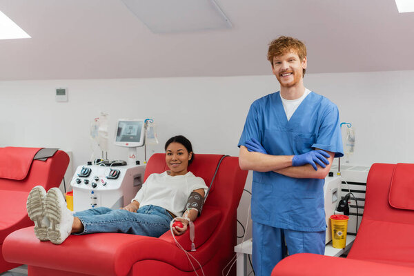happy redhead doctor in blue uniform and latex gloves looking at camera near transfusion machines and multiracial woman on medical chair donating blood in hospital