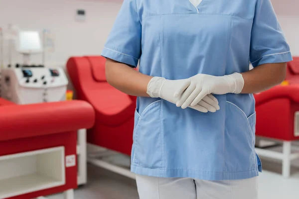 青い制服とラテックスの手袋を着た多人種間の医療従事者の一部の見解は 血液提供センターでぼやけた医療椅子や輸血機械の隣に立って — ストック写真
