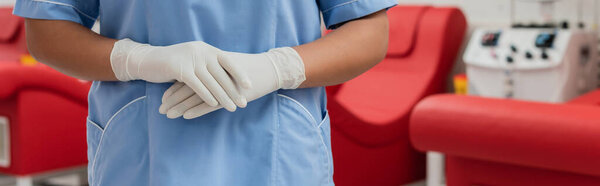 cropped view of multiracial healthcare worker in blue uniform and latex gloves standing in sterile environment of blood transfusion center, banner
