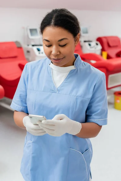 在输血中心 穿着蓝色制服和乳胶手套的多种族微笑妇女在模糊的输血机和医疗椅附近使用手机 — 图库照片