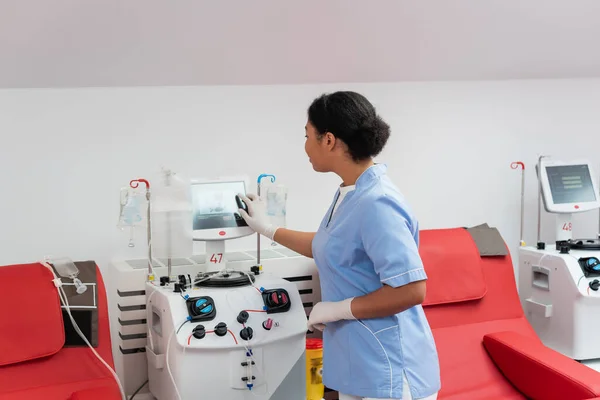 年轻的多种族医生 身穿蓝色制服 戴着乳胶手套 在献血中心靠近医疗椅的地方操作输血机 触摸屏 — 图库照片