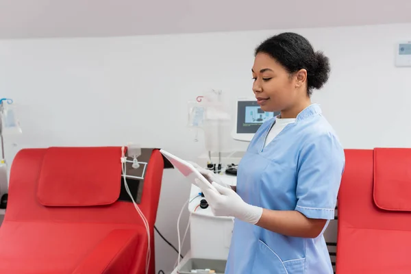 在献血中心 身穿蓝色制服和乳胶手套的多种族保健工作者在医疗椅附近使用数码平板和输血机 — 图库照片