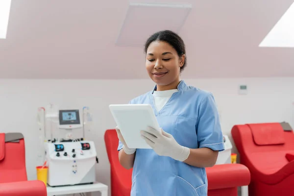 在输血站 身穿蓝色制服和乳胶手套的多种族微笑妇女在自动输血机和医疗椅附近使用数码平板电脑 — 图库照片
