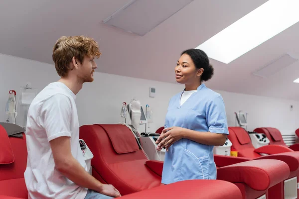 身穿蓝色制服的多种族保健工作者微笑着与坐在输血中心的医疗椅上的红头发男子交谈 — 图库照片