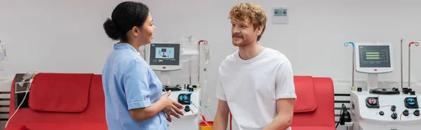 stock image multiracial nurse in blue uniform talking to happy redhead volunteer sitting on medical chair near transfusion machines in blood donation center, banner