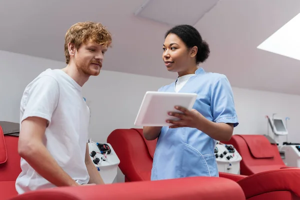 stock image multiracial nurse in blue uniform showing digital tablet to happy redhead blood donor sitting on comfortable medical chair near automated transfusion machine in hospital