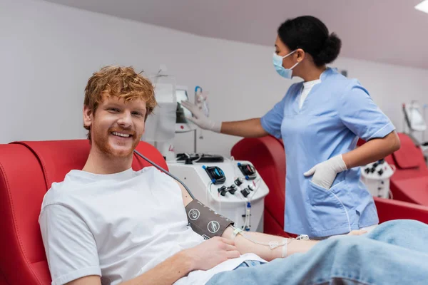 戴医疗面罩和乳胶手套的多种族护士在快乐红头发男子身边操作输血机 在献血中心看着相机 — 图库照片