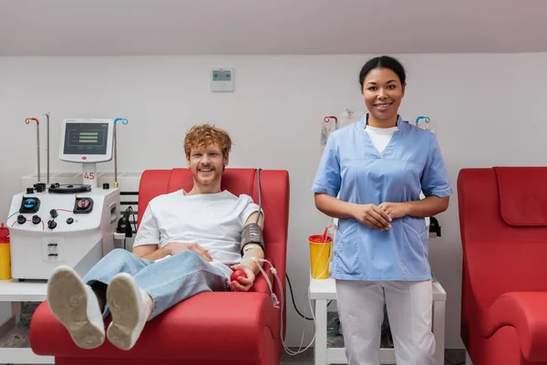 happy redhead man with blood pressure cuff and rubber ball sitting on medical chair near multiracial nurse, transfusion machine and plastic cups in laboratory, looking at camera