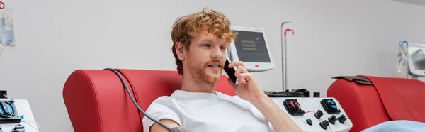 young and redhead volunteer talking on mobile phone while donating blood on medical chair near modern automated transfusion machine in laboratory, banner