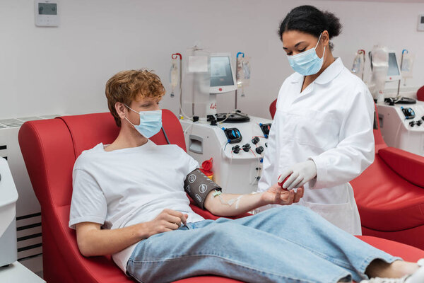 multiracial doctor in medical mask giving rubber ball to redhead donor in blood pressure cuff sitting on medical chair near transfusion machine in blood donation center
