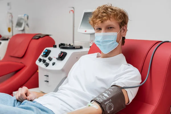 医院自动输血机附近的人体工程学座椅上 戴着医用面罩 戴着血压计的红头发捐献者坐在椅子上看着相机 — 图库照片