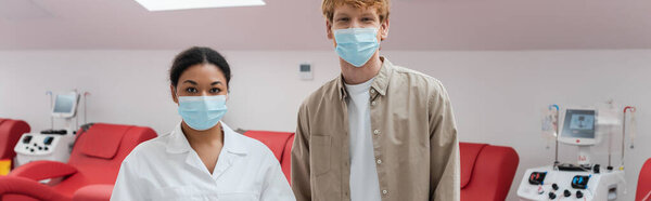 многорасовый врач и рыжий волонтер в медицинских масках, смотрящий в камеру возле аппаратов для переливания крови и медицинских стульев в центре донорства крови, баннер