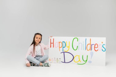 Günlük giysiler içinde gülümseyen genç kız gri arka planda, mutlu çocuk günü mektuplarıyla birlikte plaketin yanında otururken kameraya bakıyor.