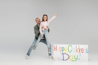 Full length of cheerful father in casual clothes holding preteen daughter on hands near placard with happy children's day lettering during celebration on grey background clipart