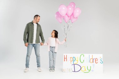 Gri arkaplanda mutlu bir çocuk günü yazısıyla neşeli balonlar ve babanın elini tutan günlük kıyafetlerle gülümseyen reşit olmayan kız.