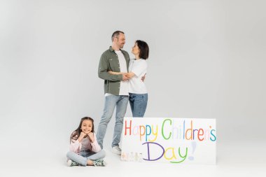 Smiling preteen girl in casual clothes looking at camera while parents hugging and standing beside placard with happy children's day lettering during celebration on grey background clipart