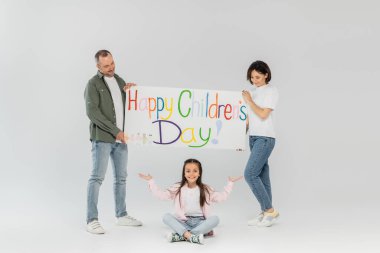 Smiling parents holding placard with happy children's day lettering and looking at preteen daughter pointing with hands during celebration in June on grey background clipart