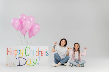 Heyecanlı ve neşeli bir anne ve günlük giysiler içinde küçük bir kız çocuğu üzerinde mutlu çocuk günü yazıları ve gri arka planda şenlik balonları olan plaketin yanındaki kameraya bakıyor.