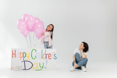 Gülümseyen, dövmeli bir anne pembe balonların yanında duran, gri arka planda mutlu çocuk günü yazılı bir pankart giymiş, günlük kıyafetli pozitif kıza bakıyor.