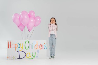 Gri arkaplanda mutlu bir çocuk günü yazısıyla pembe balonlar ve afişin yanında kameraya bakarken günlük kıyafetlerini giymiş kaygısız bir genç kız.
