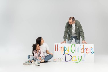 Gülümseyen kadın ve reşit olmayan kız babaya bakıyor. Mutlu bir çocuk günü tabelasının yanında duruyor. Haziran ayındaki kutlamada gri arka planda fotokopi alanı ile birlikte.
