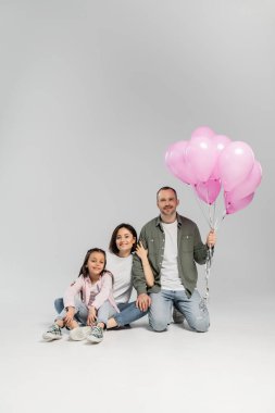 Neşeli ebeveynler ve pembe balonlarla çocuk koruma gününü kutlayan reşit olmayan kızlar ve gri arka planda birlikte kameraya bakıyorlar.