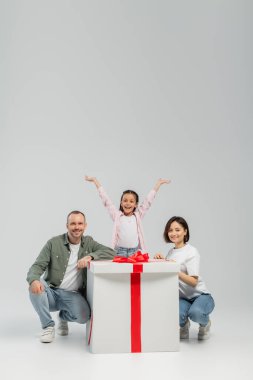 Gri arka planda uluslararası çocuk koruma günü kutlamalarında ebeveynlerin yanında duran gülümseyen çocuk ve büyük hediye kutusu.