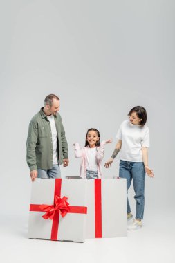 Full length of cheerful parents looking at preteen daughter looking at camera while standing inside of big present box during child protection day on grey background clipart