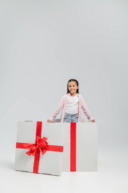 Gri arka planda çocuk koruma günü kutlaması sırasında büyük hediye kutusunda dikilirken kameraya bakan kaygısız çocuk.